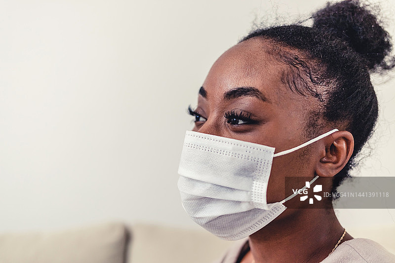 在冠状病毒/COVID-19大流行期间，美丽的非裔美国妇女戴着防护口罩坐在家里的沙发上。年轻女子戴着口罩，面带微笑。图片素材
