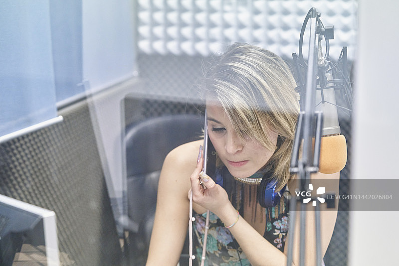 侧视图年轻的女播音员看着音频控制，而使用移动电话在电台演播室图片素材