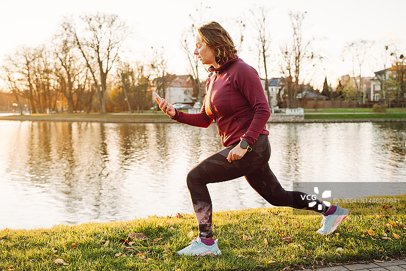 用智能应用程序晨练。女子拉伸做前弓步，在绿色公园的湖边检查智能手机图片素材