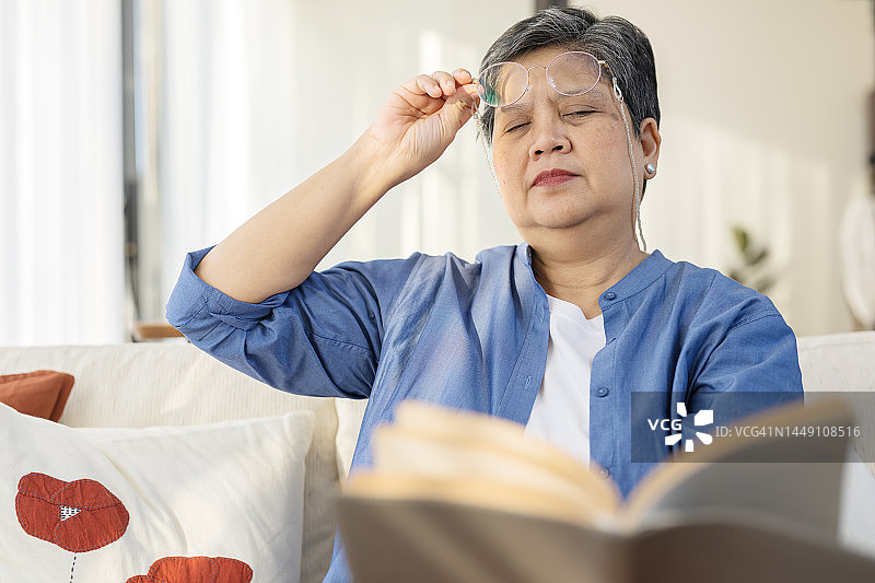 老花，由于老化导致的视力下降。一位亚洲女性在家里的客厅里看书时摘下眼镜的低角度视图。图片素材