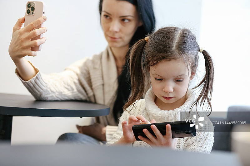 可爱的5岁女孩玩智能手机，而她年轻的成年母亲在城市咖啡馆自拍图片素材