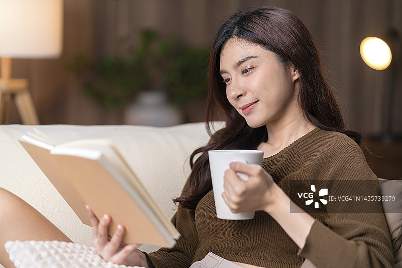 有吸引力的亚洲年轻成年女性在她家里的沙发上看书。年轻漂亮的亚洲女人在舒适的家里一边喝咖啡一边看书图片素材
