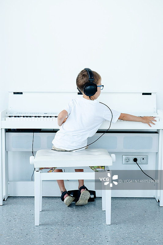一个戴着眼镜、戴着大耳机、穿着白色t恤的小男孩在音乐课上弹奏一架白色数码钢琴图片素材