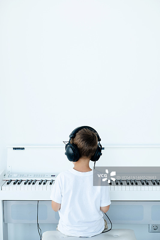 一个戴着眼镜、戴着大耳机、穿着白色t恤的小男孩在音乐课上弹奏一架白色数码钢琴图片素材
