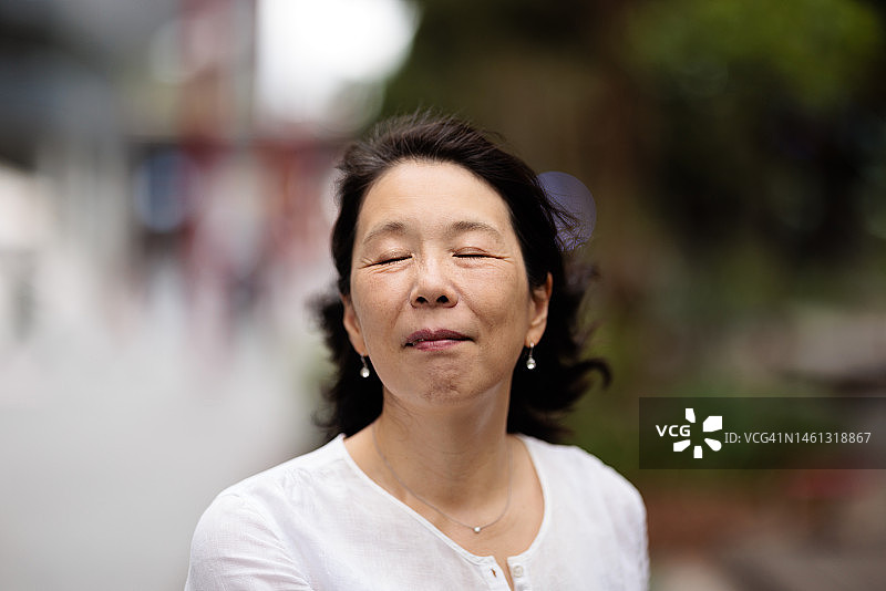 一个微笑的亚洲妇女的肖像在户外风在她的头发和她的眼睛闭着图片素材