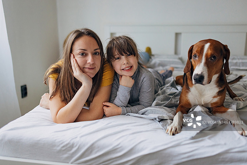 一个微笑的小女孩的肖像，她的母亲和三色狗躺在卧室舒适的床上图片素材