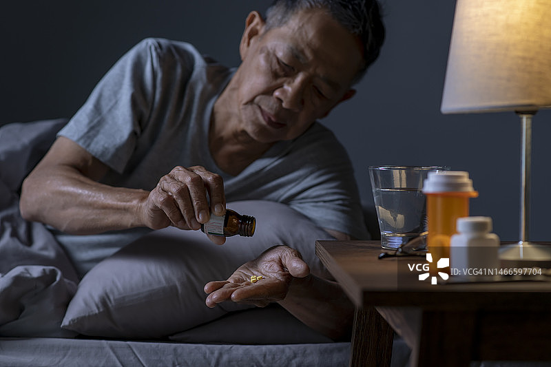抑郁的亚洲老年男子躺在床上无法入睡，因为失眠。老男人在卧室吃药。图片素材