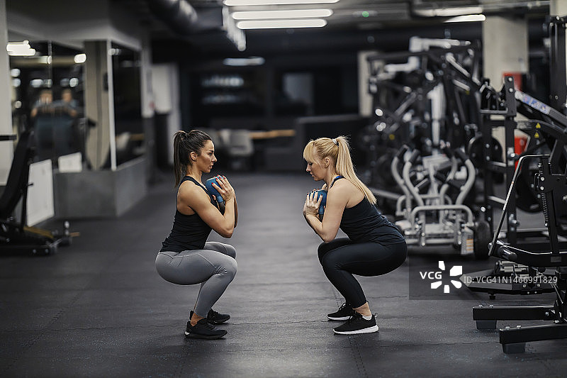 两个肌肉发达的女性朋友在健身房训练时蹲着拿壶铃。图片素材