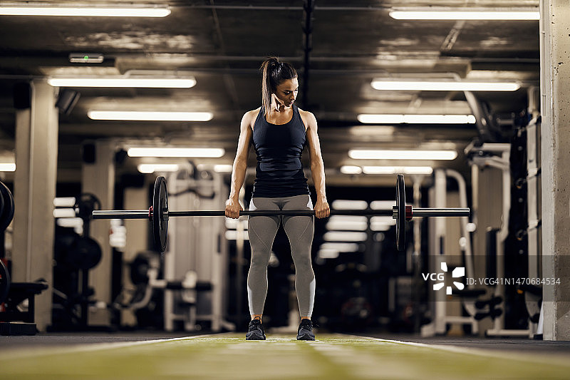 一位健康的女健身运动员正站在健身房，举着沉重的杠铃。图片素材