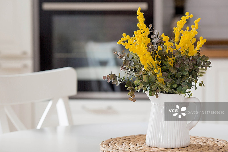 白色的桌子上放着一束含羞草。背景是斯堪的纳维亚风格的白色厨房内部。家庭舒适的概念。3月8日国际妇女节的概念。图片素材