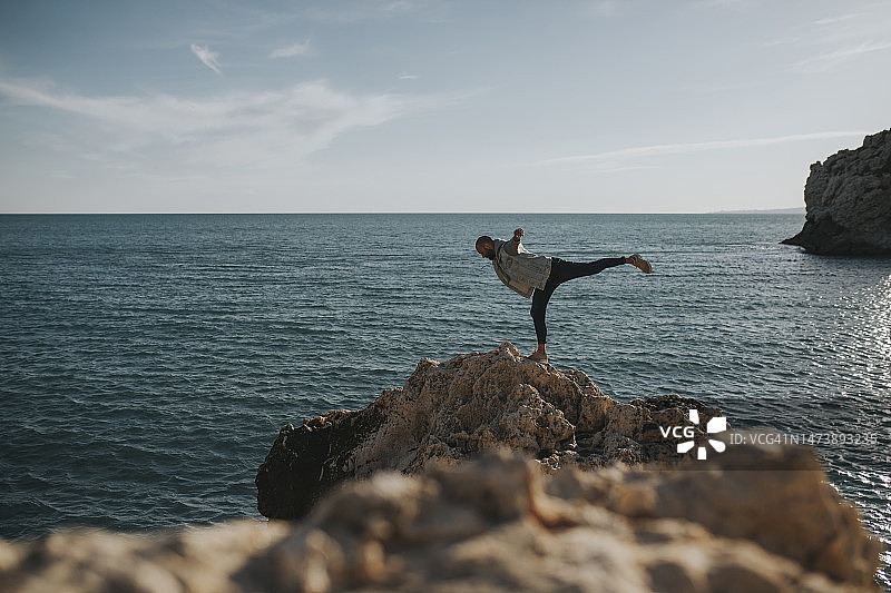 男人单腿在大海前的岩石上保持平衡图片素材