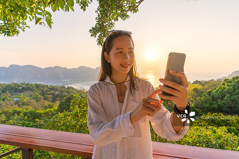 一个拿着手机的女人在皮皮岛的观景台上看山，金色的阳光照在海面和树木上。图片素材