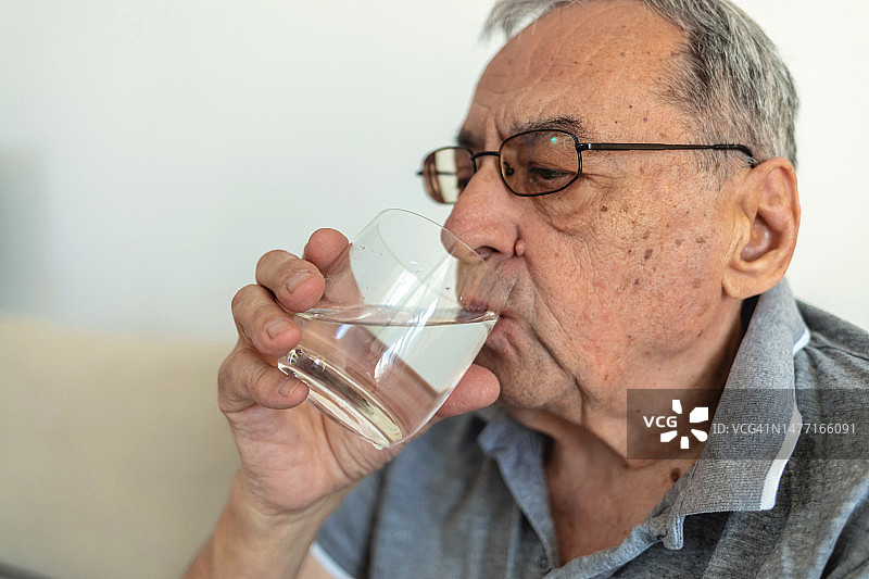 年长的白人男子坐在沙发上，拿着一杯淡水放松。白发苍苍的老爷爷在家里用水提神。图片素材