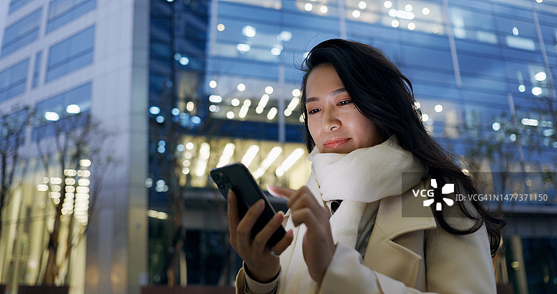 自信的年轻女子在下班的路上使用智能手机在晚上办公大楼外图片素材