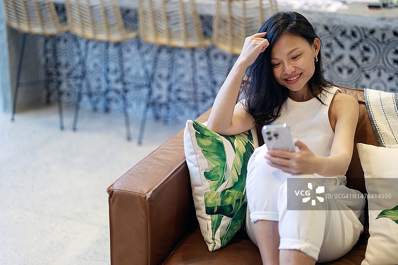 一位无忧无虑的亚洲年轻女子在家里舒适的沙发上放松地玩着智能手机。这代表了科技与日常生活的融合。图片素材