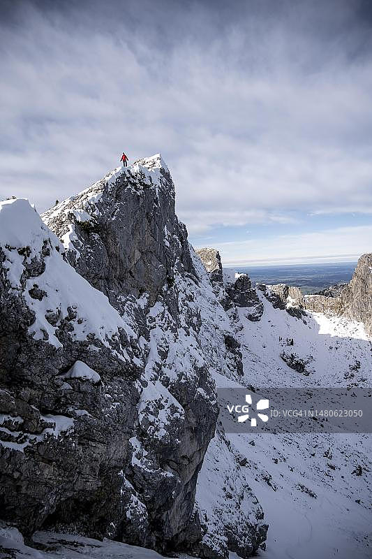 身穿红色夹克的登山运动员站在岩石塔尖上，冬天有雪的山脉，徒步前往德国巴伐利亚阿默高阿尔卑斯山的阿默高Hochplatte图片素材