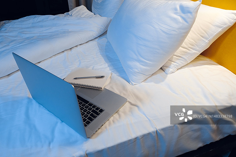 在房子或旅馆的卧室里，一间床上有一台开着的电脑的空的舒适的房间。没有人。笔记本电脑，记事本和笔在床上。周末和工作在家，休闲和生活方式。无线技术的概念，日常日常。图片素材