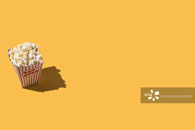 爆米花在硬纸板桶与白色和红色装饰，在左边，在黄色的背景。电影院，娱乐，电影，演出，肥胖，不健康食品，快餐和肥胖的概念。图片素材