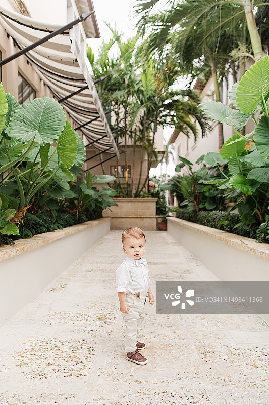 2023年5月，佛罗里达州热带棕榈滩，一个1岁的古巴裔美国小男孩，一头浅棕色短发，戴着鼠尾草绿色领结，穿着背带裤，白色纽扣衬衫，裤子和棕色鞋子，在花园里散步图片素材