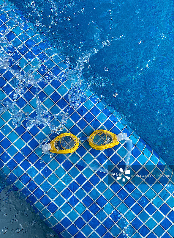 在泳池边潜水的黄色眼镜的俯视图。水在它附近飞溅。文本的负空间。图片素材