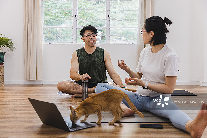 亚洲男女在练习瑜伽后使用笔记本电脑，姜黄色的猫在他们身边图片素材