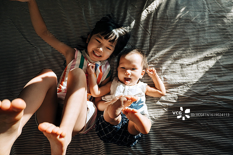 正上方的照片是快乐的亚洲小女孩和她的小妹妹在床上玩得很开心，他们俩都笑得很开心。兄弟姐妹的爱。爱，联系和团聚图片素材