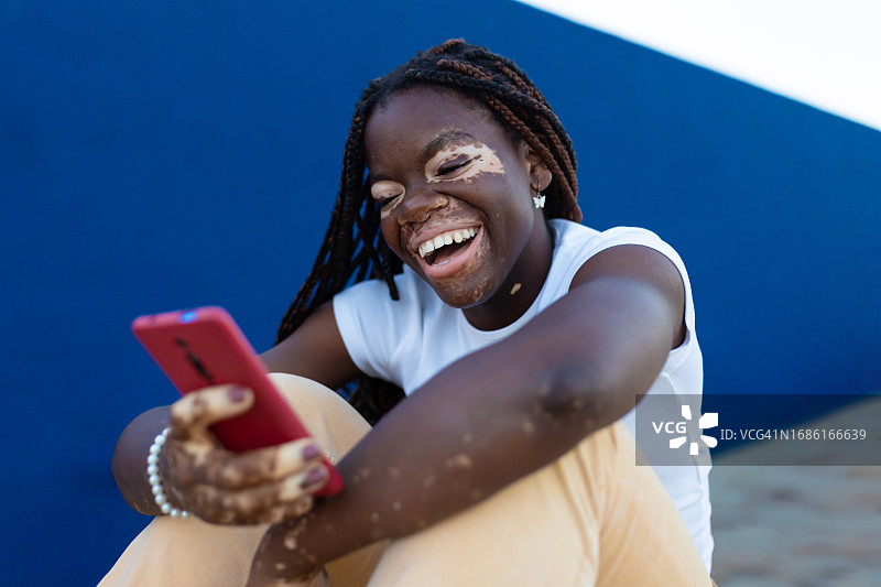 皮肤上有白癜风的快乐少女使用手机设备坐在户外蓝色背景下，感觉积极和笑。科技、青年文化和多元化的人的理念。图片素材