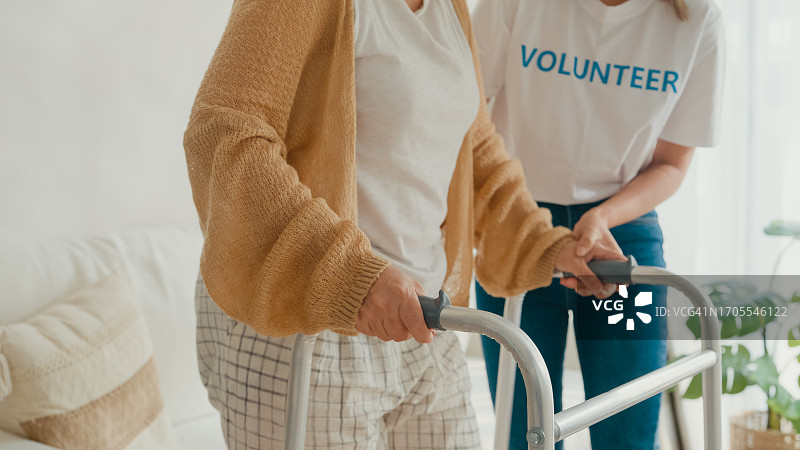 年轻的亚洲女性志愿者在家里的客厅里教老妇人走路，使用助行器照顾老人。志愿医疗保健。图片素材
