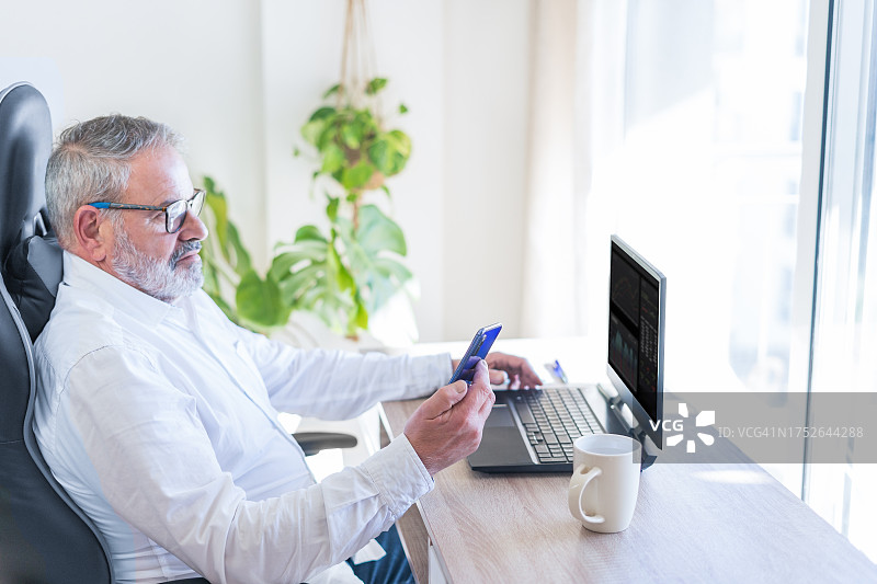 活跃的老人在办公室用笔记本电脑工作，他穿着白衬衫查看智能手机。图片素材
