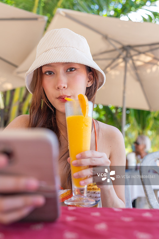 Z世代亚洲女性一边使用智能手机，一边在海滨餐厅享用芒果果汁混合饮料图片素材