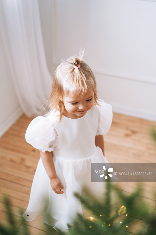 可爱的微笑的小女孩，穿着白色的裙子，在客厅的圣诞树旁图片素材