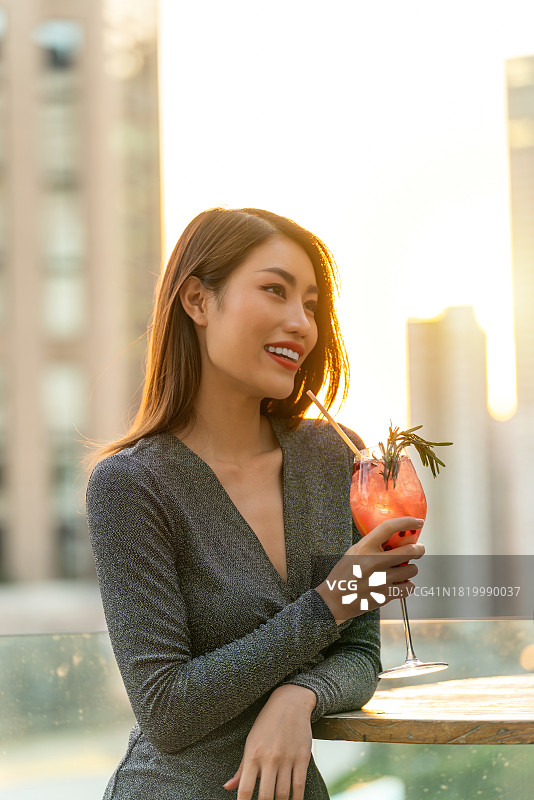 在温暖的晚霞中，一个亚洲女人在屋顶酒吧喝鸡尾酒图片素材