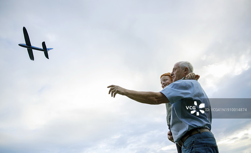 微笑的爷爷抱着孙子，在多云的天空下飞着玩具飞机图片素材