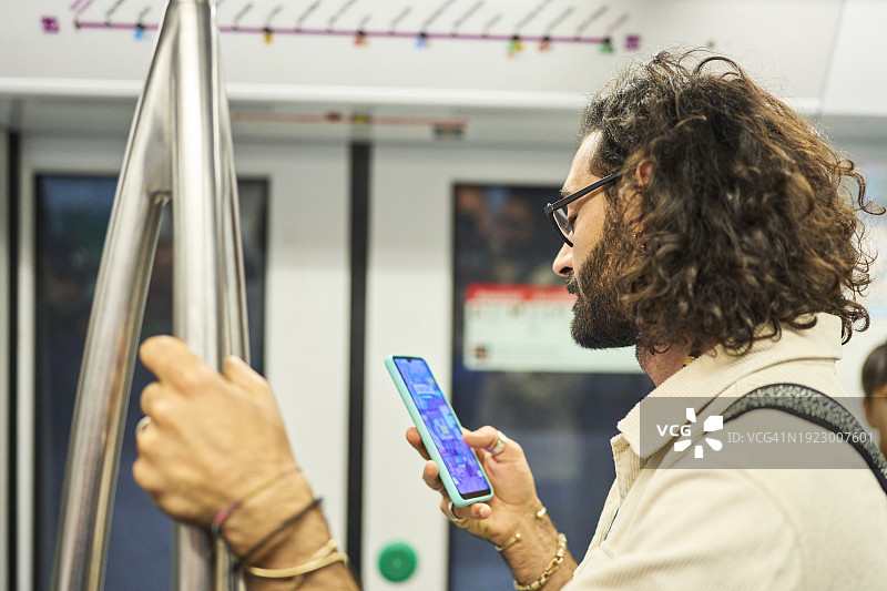 一个留着长发、戴着眼镜、留着胡子的站着的男人，在地铁里用他的智能手机。图片素材