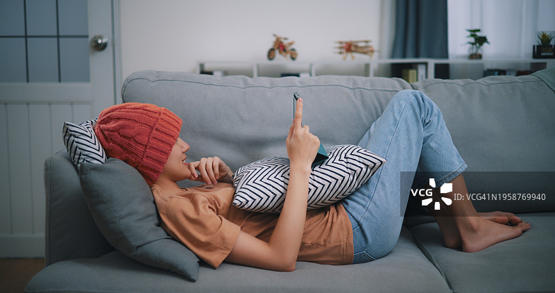 年轻的女性癌症患者躺在客厅的沙发上玩平板电脑图片素材