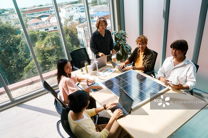 在一间现代化的办公室里，一个多元化的团队聚精会神地讨论着可再生能源，桌子上放着一块巨大的太阳能电池板。图片素材