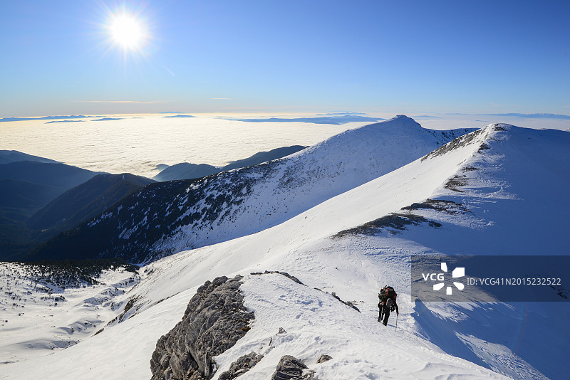 在阳光明媚的日子里，男徒步旅行者在白雪皑皑的山脊上到达山顶图片素材