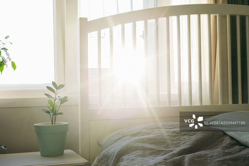 白色的床立在卧室的窗边，夕阳和一缕阳光。桌上的家庭植物图片素材