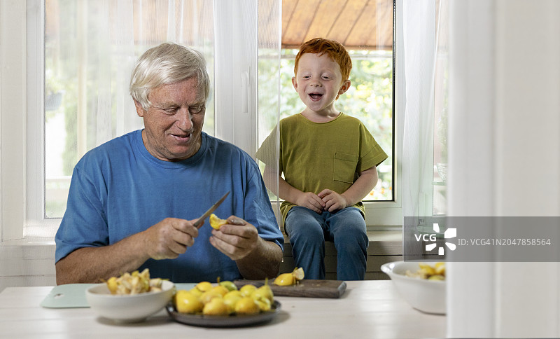 快乐的男孩坐在家里切水果的爷爷旁边图片素材