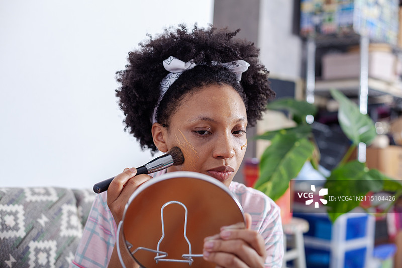 一位黑人妇女在脸上涂粉底，坐在沙发上看着一面小镜子图片素材