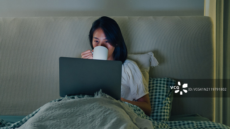 年轻的亚洲女孩晚上坐在家里的床上使用笔记本电脑和喝牛奶。失眠、晕机、无手机恐惧症、睡眠障碍。图片素材