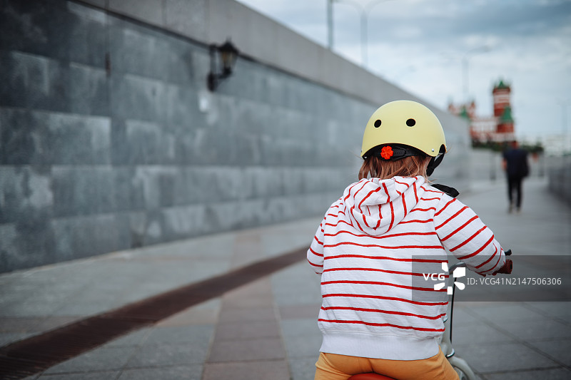 蹒跚学步的女孩骑着头盔在灰色小路上探索城市滨水区图片素材