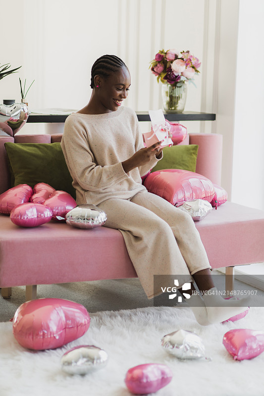 漂亮的黑人女孩坐在沙发上，围着粉红色的气球，打开礼盒。图片素材