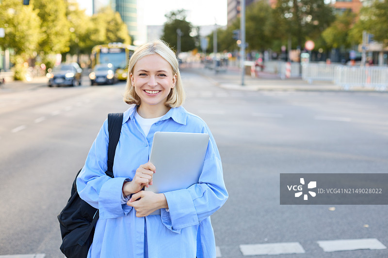 街头时尚的年轻快乐女子肖像，金发女孩背着背包和笔记本电脑，站在户外图片素材