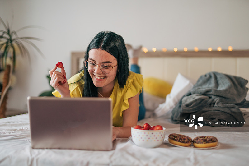 年轻女子躺在床上享受周末，用笔记本电脑看电影，吃着草莓和甜甜圈图片素材