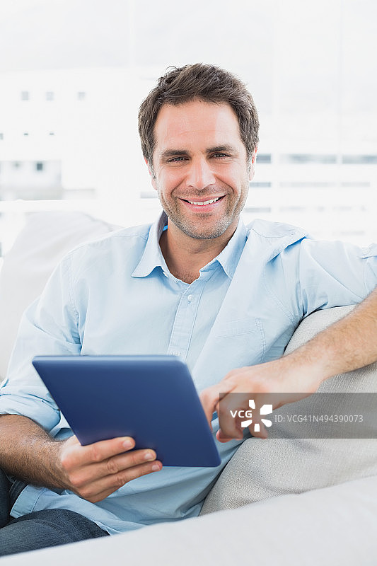 英俊的微笑男子坐在沙发上使用他的平板电脑图片素材