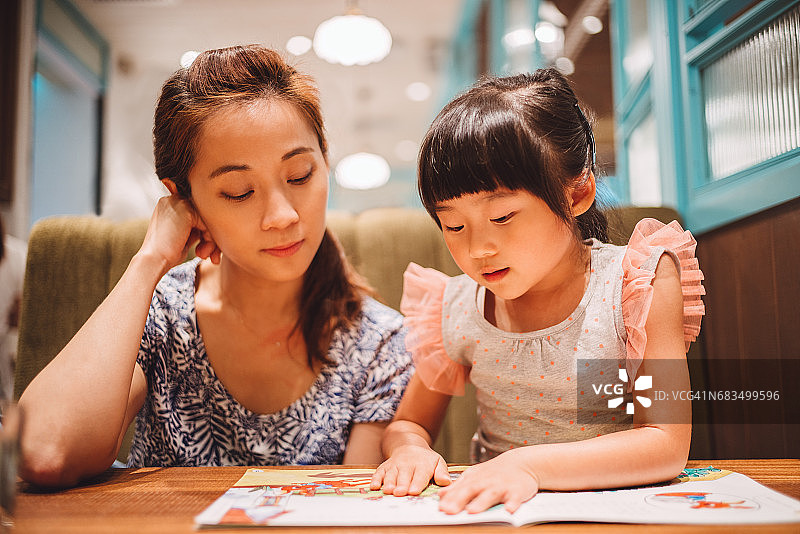 漂亮的年轻妈妈和她可爱的小女儿在一家餐馆里快乐地读着书。图片素材