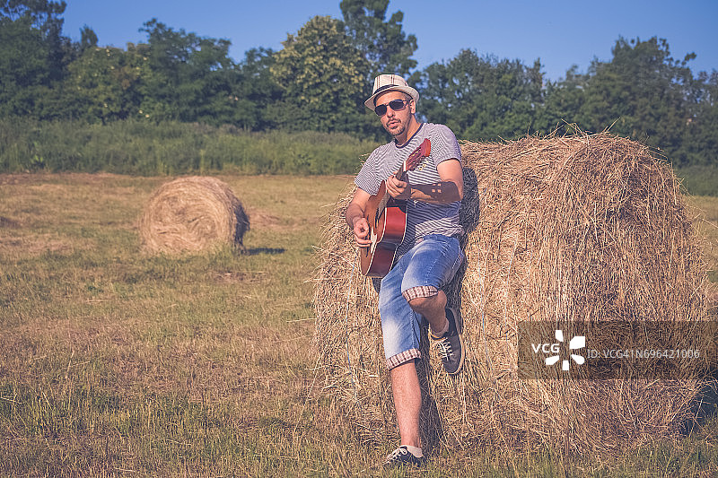 年轻时尚的音乐家与原声吉他在草地上靠在干草卷。音乐，艺术和生活方式的概念。图片素材