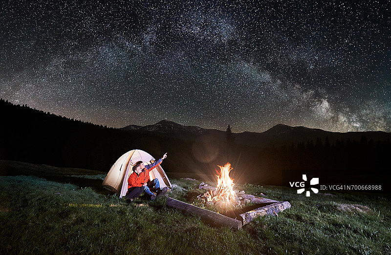 晚上在山里露营。一对游客坐在篝火旁被照亮的帐篷里，看着满是星星和银河的美丽夜空。人指着天空。长时间曝光图片素材