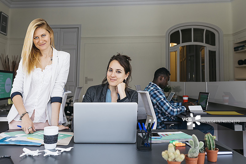 年轻的商业女性一起工作，使用笔记本电脑，同事坐在后台图片素材